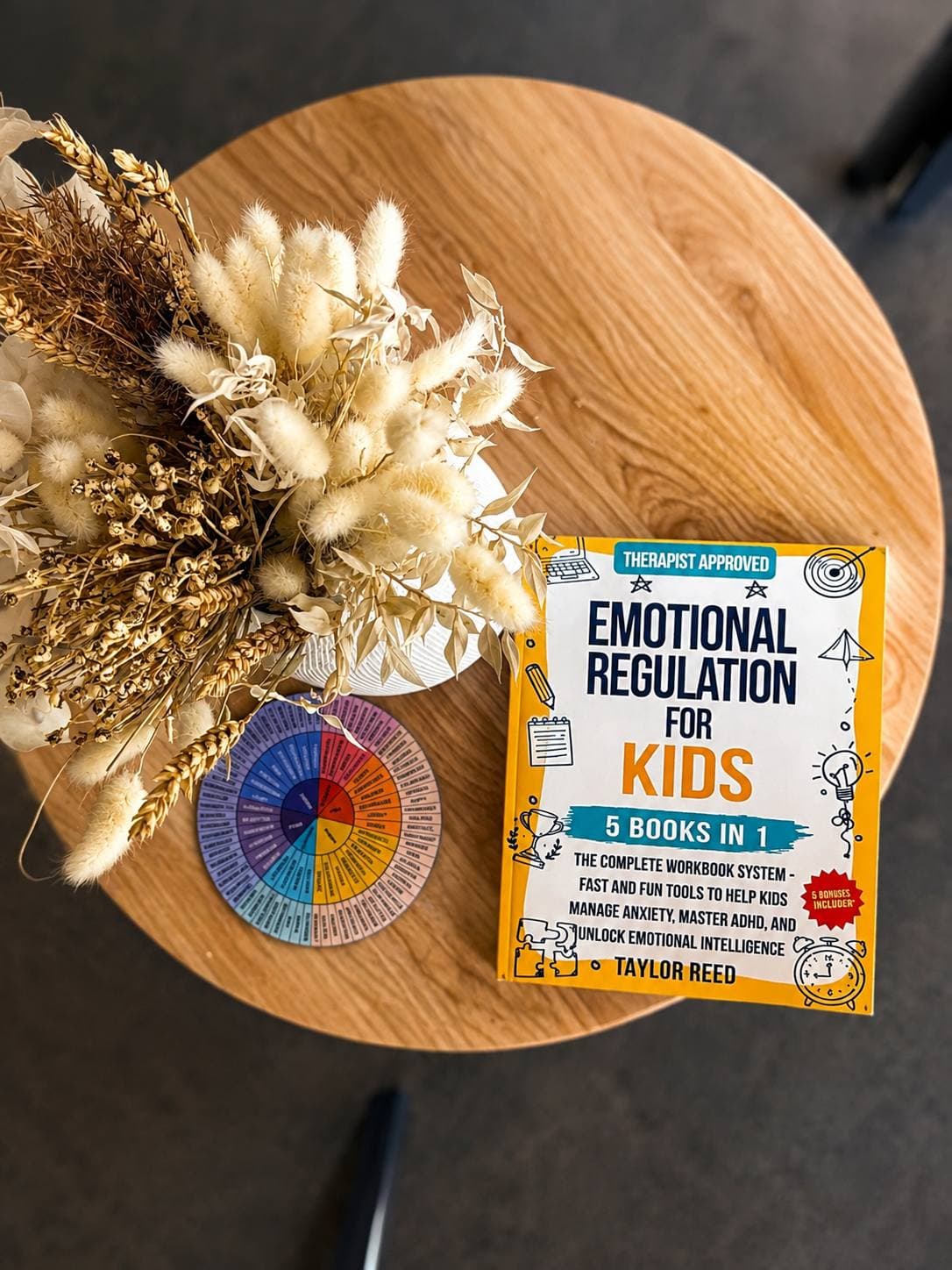 A round table with a feelings wheel, a book on emotional regulation for children and a vase of dried flowers